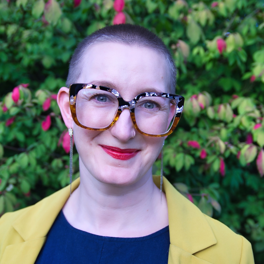 Dr. Stef Sloan wearing wide rim glasses, smiling outdoors, and surrounded by plants and pink flowers.