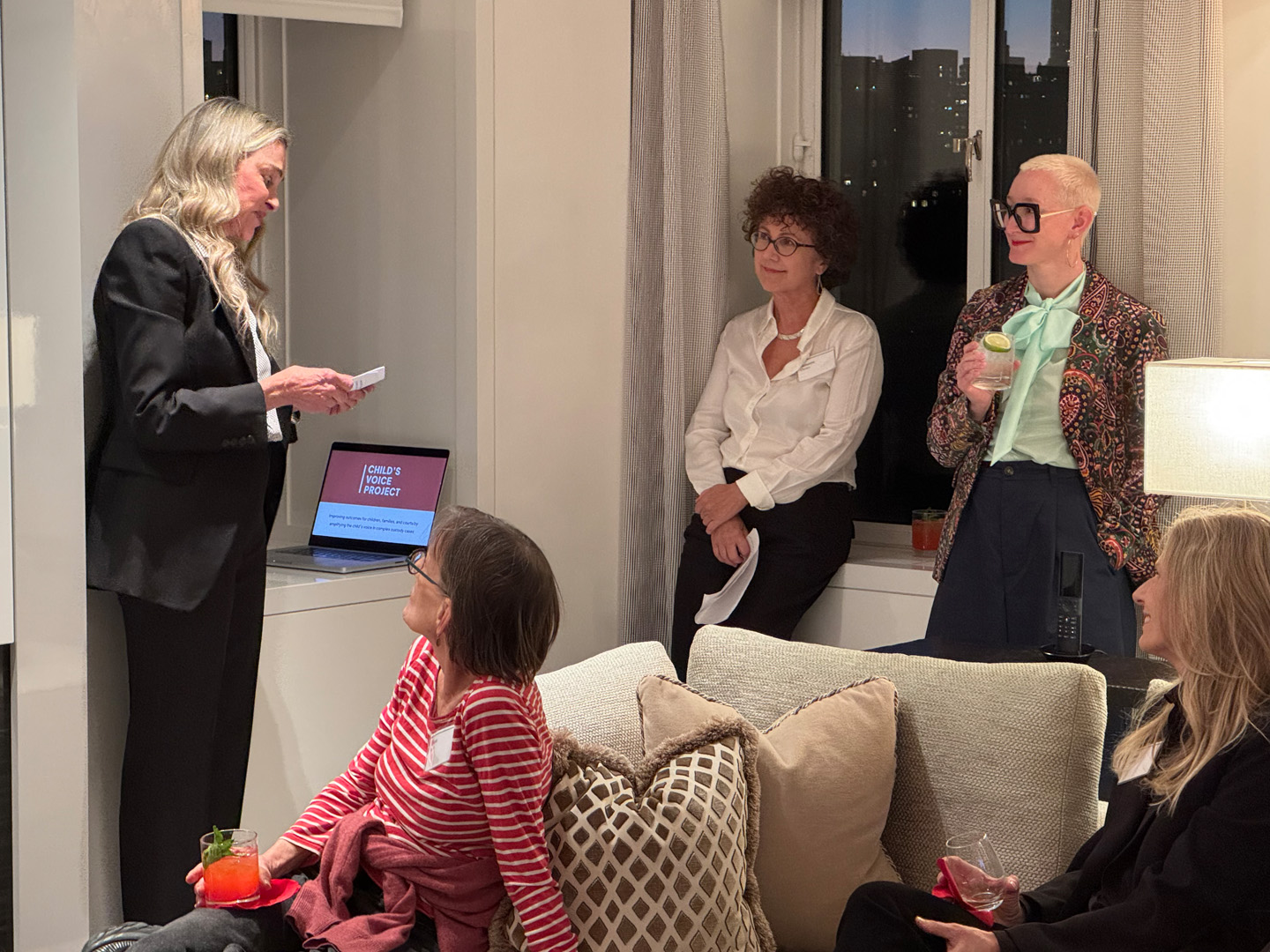 Woman in a black suit reads from a notecard near a laptop showing the “Child’s Voice Project” logo, while two other women stand nearby and a seated guest listens.