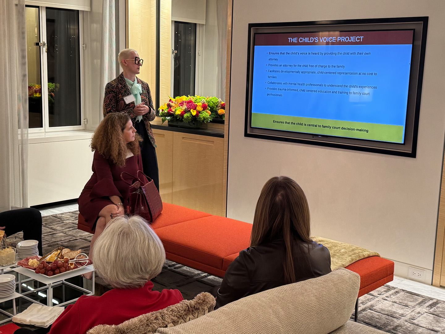 Speaker presenting slides about “The Child’s Voice Project” on a large screen as guests seated nearby listen and observe.